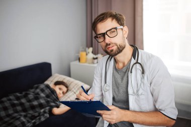 Ciddi genç erkek doktor bak gözlük sayesinde fotoğraf makinesinde. Yatakta yatarken onun ptient. Küçük kız battaniye ile örtülü. O hasta.