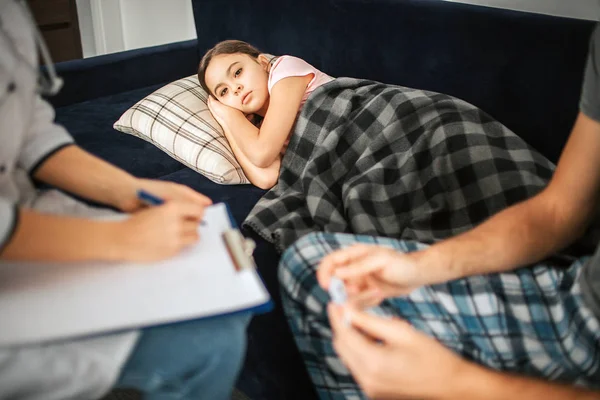 Bild eines kleinen Mädchens auf dem Sofa liegend. Sie bedeckte sich mit einer Decke. Ärztin und Vater sitzen neben dem Mädchen. er hält Thermometer. Arzt schreibt Medikamentenverschreibung. — Stockfoto