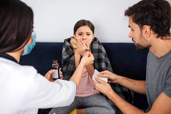 Verängstigte und verängstigte Mädchen schauen in die Kamera. Sie bedeckt den Mund mit Fingern. Ärztin gibt ihren Löffel mit Sirup-Medizin. Vater hält weißes Gewebe. — Stockfoto