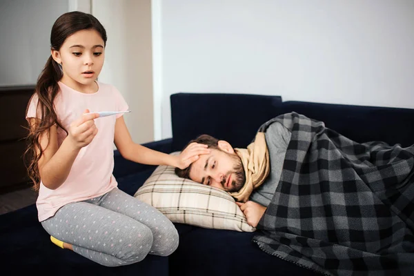 Una niña asustada sentada junto a su padre enfermo. Ella mira el termómetro y se pregunta. Joven, duerme. Está enfermo y tiene fiebre. . — Foto de Stock