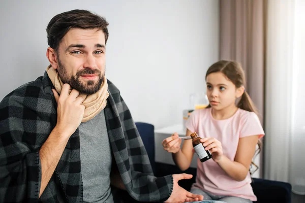 Bild eines jungen Mannes, der mit seiner Tochter auf dem Sofa sitzt. er schaut in die Kamera und hält die Hand an der Kehle. Es ist mit einem Schal bedeckt. Decke auf seinen Schultern. Mädchen gibt ihm Behandlungssirup. — Stockfoto