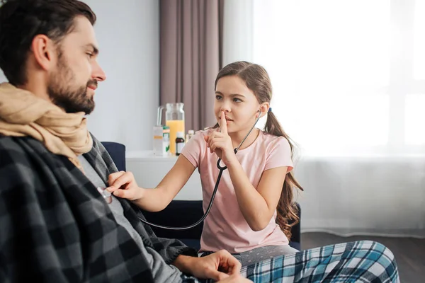 Small girl listen to dads breathing through stethoscope. She hold finger on lips and look at him. Sick young man covered with blanket and scarf.