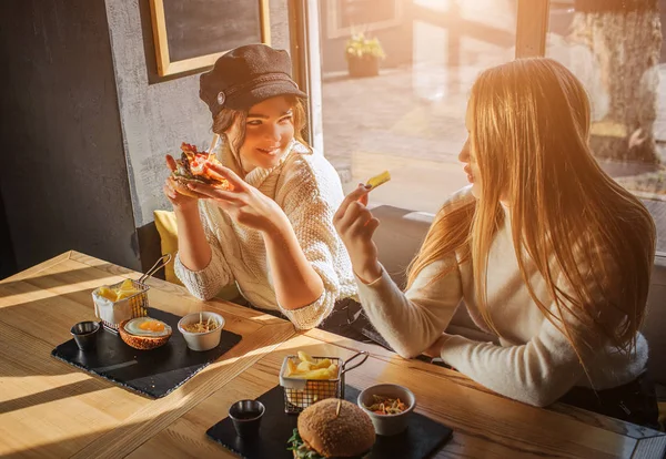 Счастливые молодые женщины сидят за столом и смотрят друг на друга. Модель фирмы держит бургер с беконом. Во втором кусочек Санни снаружи. . — стоковое фото