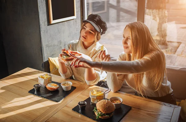 Twee jonge vrouwen zitten in op tabe. Model op het juiste punt naar voren. Ten tweede houdt een hamburger in de hand. Ze kijken uit. — Stockfoto