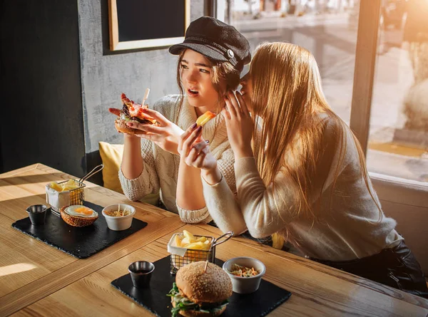 Jovens mulheres bonitas sentam-se ao lado da mesa. Eles fofocam. Blodne menina wisper no ouvido para outra menina. O modelo na tampa é espantado. Ela segura hambúrguer. . — Fotografia de Stock