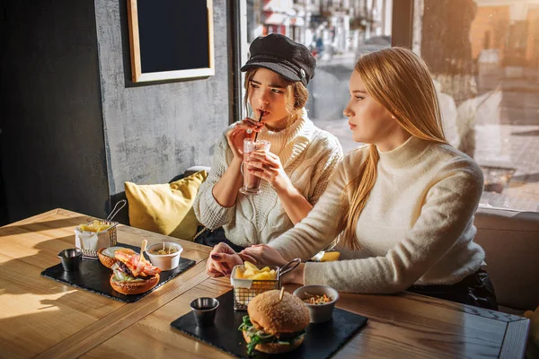 Twee mooie tieners zitten aan tafel in een cafe en kijken naar links. Eerst drink cocktail. Ten tweede zitten naast vriend. Ze zijn aat venster. Zon shiines buiten. — Stockfoto