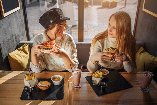 Весёлые молодые женщины смотрят друг на друга и улыбаются. Они сидят за столом в кафе и держат бургеры. Молодые женщины счастливы . — стоковое фото