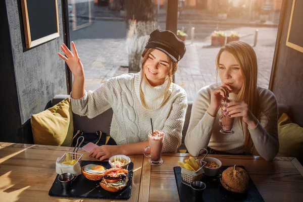 De jonge vrouw Cheeerful in de dop zit aan tafel met haar vriend. Ze zwaait met hand en glimlach. Tweede model cocktail drinken. Ze hebben voedsel aan tafel. — Stockfoto