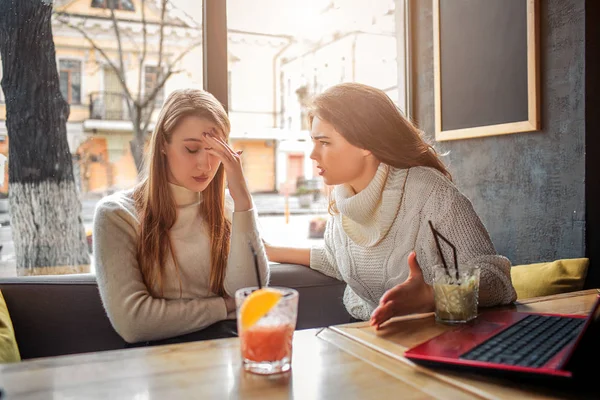 Eine aufgebrachte junge Frau sitzt am Tisch. Sie verdeckt das Gesicht mit der Hand und schaut nach unten. ihre Freundin schaut sie an und redet. — Stockfoto