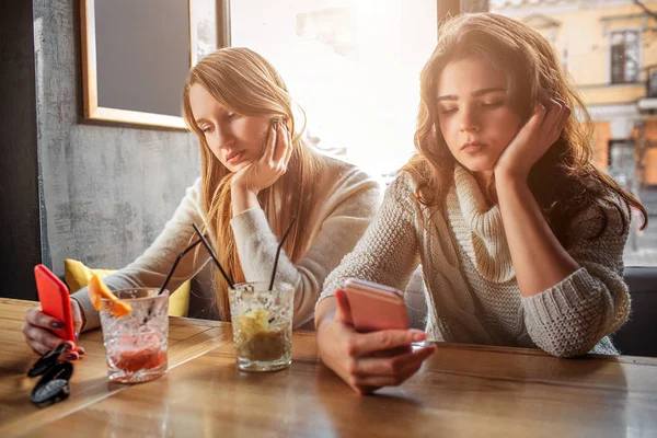 Набридли молоді жінки сидять за столом. Вони тримають телефони і дивляться на нього. Моделі мають окуляри з напоєм за столом . — стокове фото