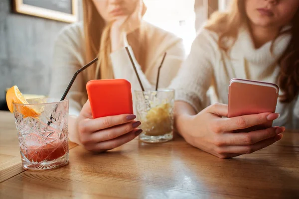 Geknipte weergave van jonge vrouwen zitten aan tafel. Ze houd de telefoon in handen. Modellen zijn vervelen. Twee glazen met cocktails worden aan tafel. — Stockfoto