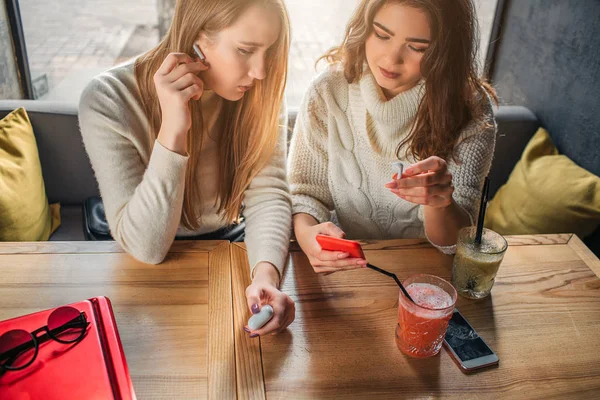 Am Tisch sitzen junge Frauen. Brünett zeigt auf das Telefon, das sie in der Hand hält. Blondine hält Finger auf drahtlose Kopfhörer. Sie sind konzentriert. Modell für rechtes Gerede. — Stockfoto