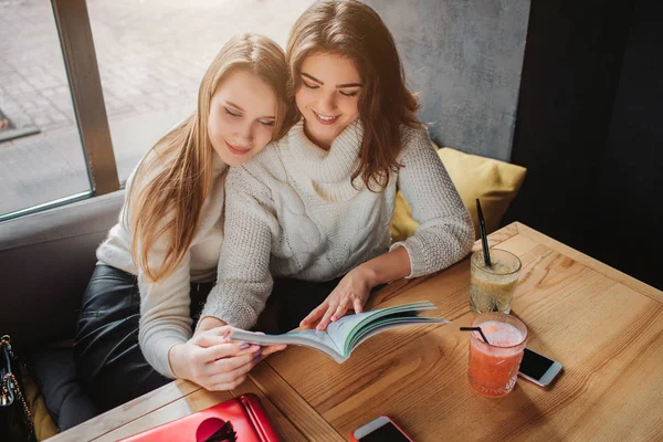 Позитивные и милые молодые женщины сидят за столом и смотрят журнал. Они улыбаются. Девушки сидят и обнимаются . — стоковое фото