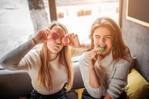 Lustige junge Frauen bedecken die Augen mit Macarons. posieren sie vor der Kamera. Brünette beißt Stück Makarone. Sie haben Spaß. — Stockfoto