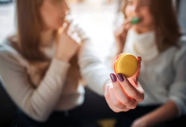 Feche a mão segurando macaron amarelo na mão. As mulheres jovens comem cookis e olham um para o outro. Sentam-se juntos. . — Fotografia de Stock