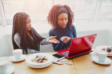Genç Afro-Amerikan kadınlar masaya oturabiliriz. Onlar laptop noktası. Hayal kırıklığına uğramış ve hayret modeller. Onlar masada yiyecek ve içecek var.