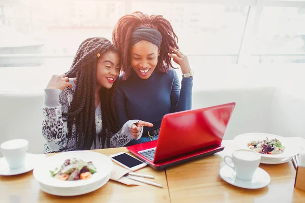 Mujeres africanas jóvenes y positivas se sientan a la mesa. Miran en el portátil. Modelos sonrían. Una mujer apuntando a la laptop. Se sientan en la cafetería . — Foto de Stock