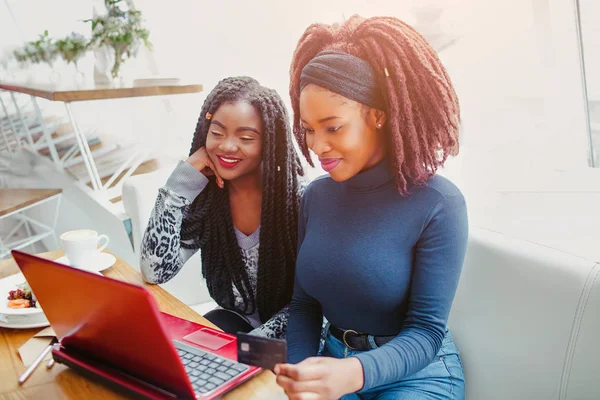 Neşeli genç Afrikalı kadınlar masaya oturabiliriz. Dizüstü bilgisayar ekranında görünüyorlar. Modelleri gülümse. Onlar kafede oturup.