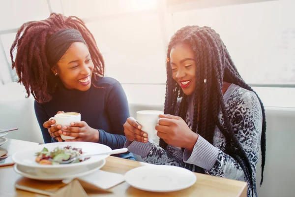 Mujeres africanas jóvenes agradables y positivas riéndose. Se sientan en el café a la mesa. Los modelos tienen tazas con café en las manos. Ellos disfrutan . — Foto de Stock