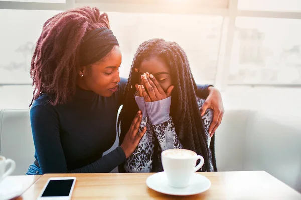 Upset youn african woman sit and cry. She covers face with hands. Her friend comfort young woman. They sit in cafe at table.