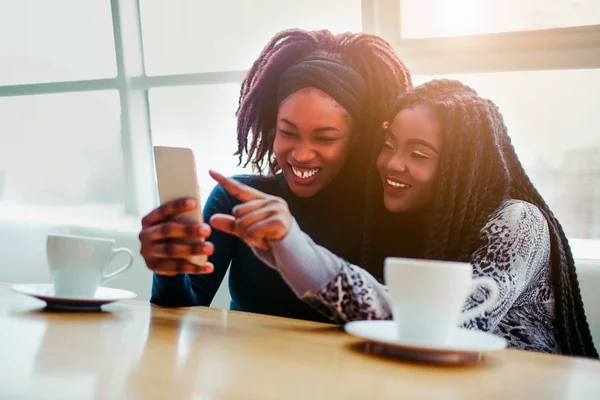Alegres jóvenes africanas se sientan juntas. Sonríen y señalan el teléfono blanco. Modelos sentarse ar bringht ventana . — Foto de Stock