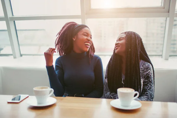 Positieve jonge Afrikaanse vrouwen lachen. Ze kijken naar elkaar en aanraking van haar. Mensen zitten op de witte Bank in de buurt van venster. — Stockfoto