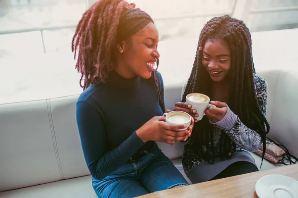 Atractivas mujeres africanas sonríen. Se sientan en la cafetería y sostienen tazas de café. Segundo modelo mira hacia abajo . — Foto de Stock