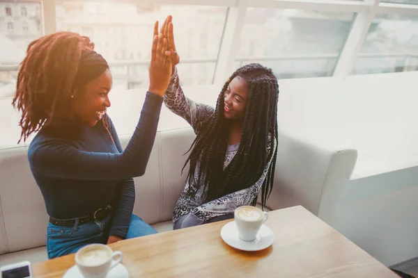 Tow Afrikaanse modellen geven hoge vijf ene naar de andere. Ze kijken naar elkaar en glimlach. Frieds doorbrengen tijd in café ter venster. — Stockfoto