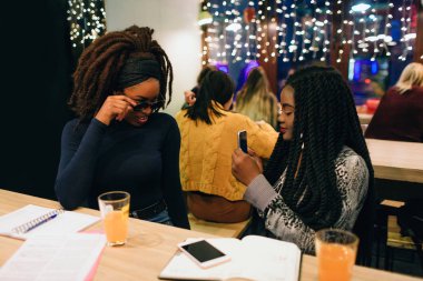 Bir Afrikalı model telefon kameraya poz. Sungalsses üzerinde elini tut. İkinci model telefon tutun ve fotoğrafı çekin. Kafede masada oturuyorlar.