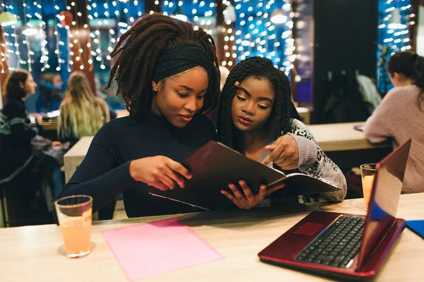 Dois estudantes africanos olham para o diário aberto. Sentam-se e estudam. Eles têm laptop vermelho na mesa . — Fotografia de Stock