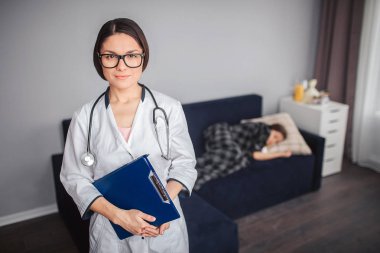 Genç kadın doktor odasında stand ve kameraya bak. Plastice tablet el ve boynuna stetoskop tutun. Küçük kız hasta uyku kanepede.