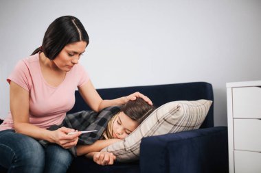 Endişeli genç kadın childs kanepede oturup termometre el bakmak. O kızı saç dokun. Kanepede kız hasta uyku.