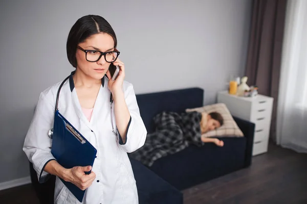 Serius preocupado jovem falar ao telefone. Ela está no quarto. A paciente dela deitada no sofá atrás. Pequena menina doente dormir . — Fotografia de Stock