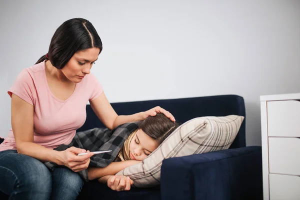 Worried young woman sit at childs couch and look at thermometer in hand. She touch hair of daughter. Sick girl sleep on couch.
