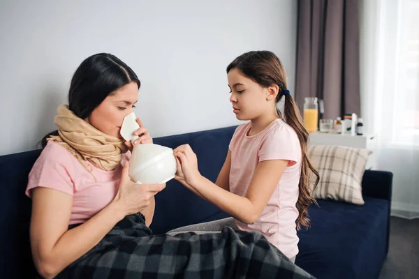 Fiatal beteg nő ül a kanapén szobában lányával. Ő lélegezzük be a gyógyszer inhalációs keresztül. Barna tart. Gyermek mellett ülni, és segíteni neki anyja. — Stock Fotó