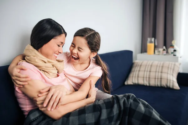 Счастливая и позитивная молодая женщина сидит вместе со своей дочерью на диване в комнате. Они смеются вслух. Молодая женщина больна. Её горло завернуто в шарф. Девушка обнимает мать . — стоковое фото