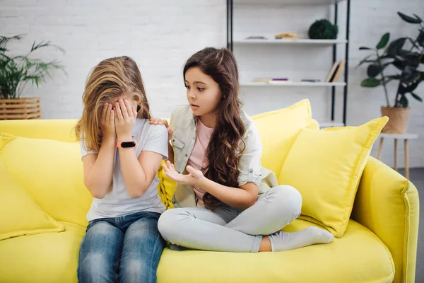 Upset girl on left hide face with hands. She cry. Teenager sit with her friend on yellow sofa in room. Brunette try to comfort her. — Stock Photo, Image