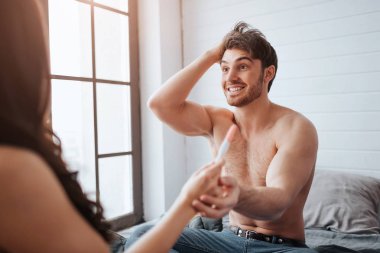 Happy young man smiling to woman. They sit on bed in room at window. Guy cheering. He hold womans hand with positive pregnancy test result.