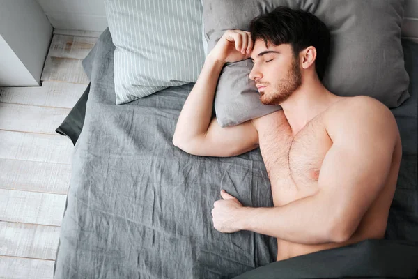 Одномісний молодий м'язистий чоловік спить у ліжку. Він тримає голову на подушці. Молодий чоловік голий. Нижня частина тіла покрита сірою ковдрою . — стокове фото