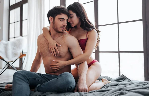 Good-looking hot couple sit on bed in room. Sexy young woman in lingerie embrace man from behind. He look at her. Satisfaction and seduction.