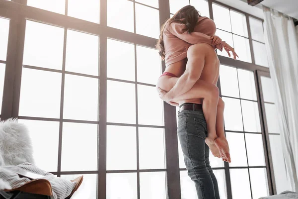 Sexy paar staan op venster en verbergen hoofd onder trui. Guy houden vrouw op handen. Ze zit op hem en omarmt zijn lichaam met benen. — Stockfoto