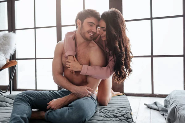 Felice e positivo giovane coppia siedono insieme sul pavimento in camera. Sta dietro e abbraccia l'uomo. Lui la guarda e sorride. Donna magra a ragazzo . — Foto Stock