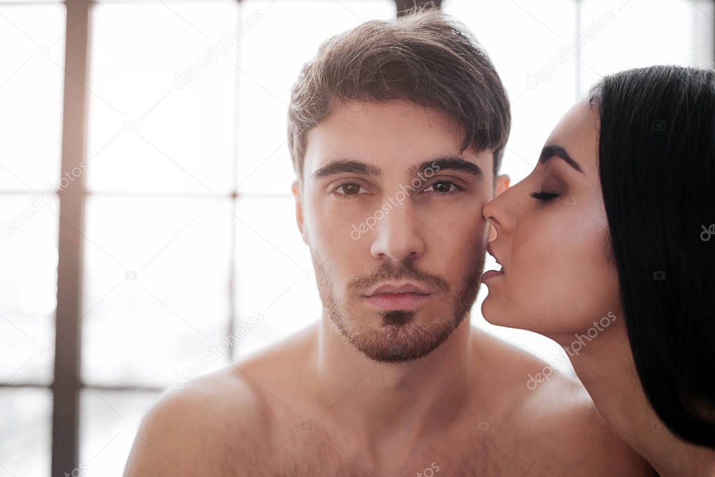 Young handsome man look on camera. Beautiful woman kiss him. Window is behind them. They are naked in room.