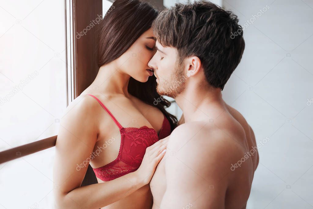 Passionate young woman in red lingerie stand at window and kiss with man. He lean to her. Model touch her partner with hand.