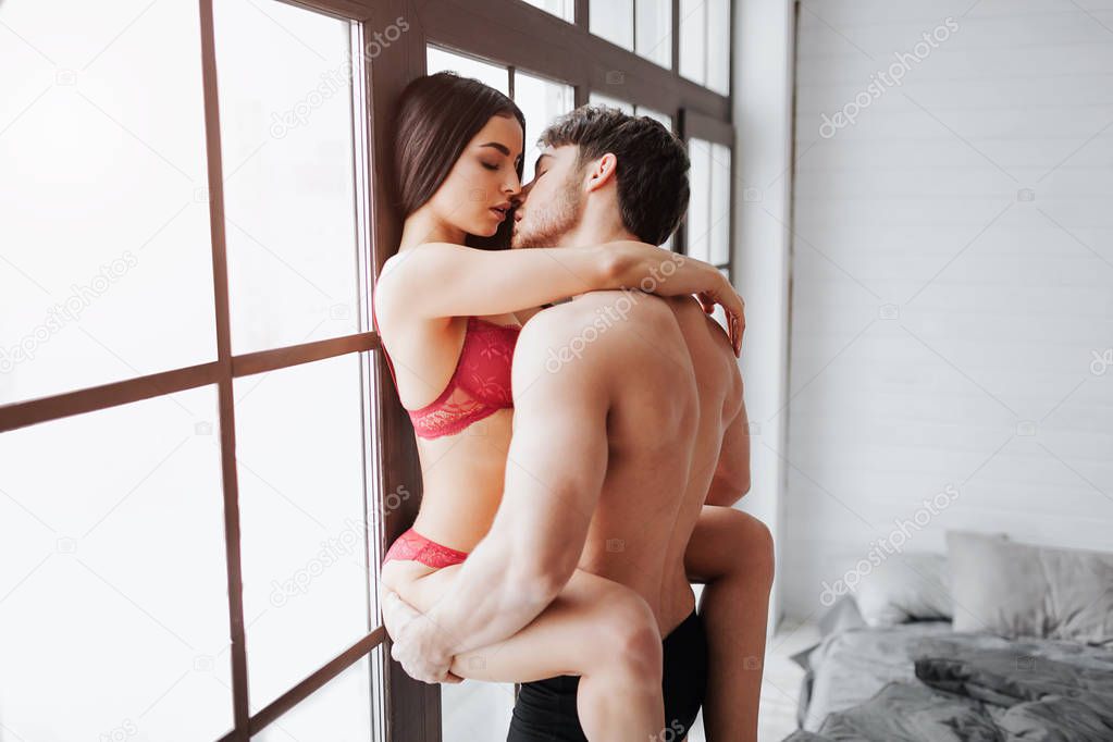 Hot young woman in red lingerie sit on man and kiss him. She embrace him with legs and hands. Guy hold woman at window. Man has hands on models buttocks.