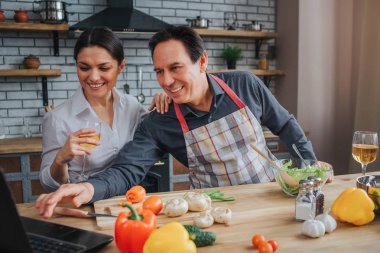 Neşeli adam karısı ile birlikte masaya otur. Gülümse ve dizüstü ekranda izlemek. Adam dokunmatik klavye. Kadın şarap tutun. Masanın üzerinde yatan sebze.