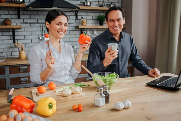 Gut gelaunte Männer und Frauen sitzen in der Küche und blicken direkt in die Kamera. sie lächeln. Frau hält Tomate auf Gabel und Paprika in einer anderen Hand. Der Mensch hat Becher. sie sitzen am Tisch. — Stockfoto