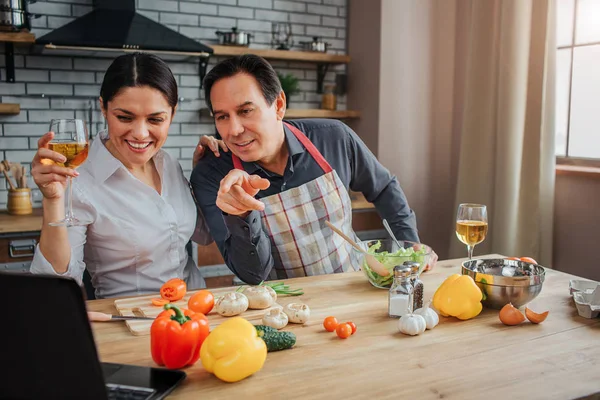 Nice couple sit at table in kitchen and watch on laptop. They hold glasses of wine and smile. Guy point on screen. They wear aprons. Couple has vegetables on table.