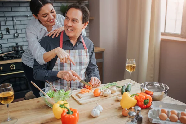 Helpful woman stand behind man in kitchen. He sit at table and look at her. They smile to each other. Guy cut tomatoes.