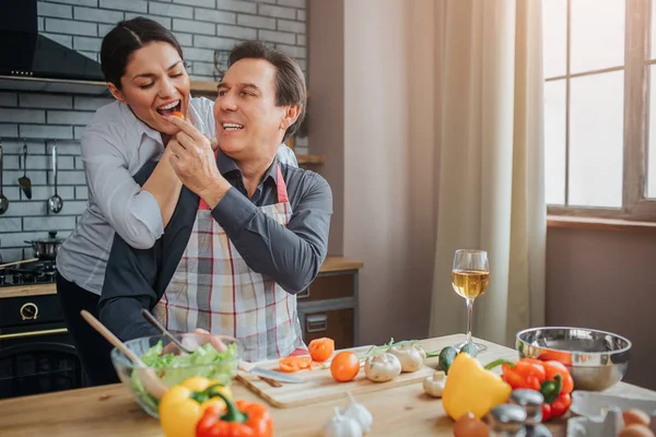 Romantic picture of man feeding woman with piece of vegetable. They are together in kitchen. He sit at table while she stand behind him. They have vegetables and glasses of wine at table.